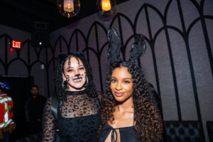 Two women in Halloween costumes posing at The Standard in Fresno. The woman on the left is dressed as a cat, wearing a black leopard-print top, cat-ear headband, and painted whiskers. The woman on the right is dressed as a bunny, wearing a low-cut black top and large black bunny ears.