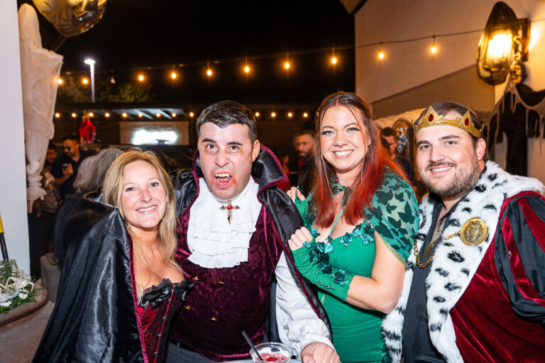 A group of four adults in themed Halloween costumes posing together at The Standard in Fresno Halloween party. The two people on the left are dressed as vampires, with the man in a burgundy velvet jacket, ruffled shirt, and exposed fangs, and the woman in a black cape. The two people on the right are dressed as royalty and a nature-themed character: the woman is wearing a green dress and a leaf-patterned cape (possibly Poison Ivy), and the man is wearing a king's costume with a red velvet and fur-trimmed cape and a gold crown. The background is an outdoor patio area lit by string lights.