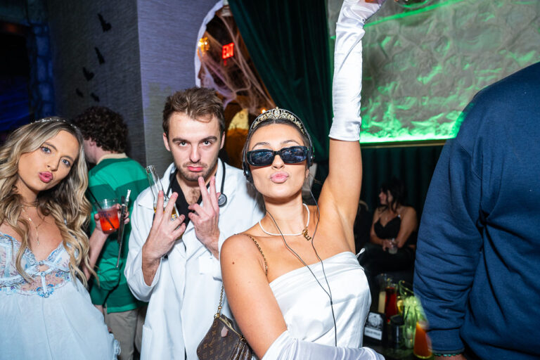 Three people in Halloween costumes posing for the camera at The Standard in Fresno Halloween party. In the foreground, a woman dressed in a white satin dress, long white gloves, and sunglasses with a tiara is raising one arm and making a kissing face. Next to her, a man is dressed as a doctor in a white lab coat with a stethoscope and is making a peace sign. On the far left, a woman in a light blue or white dress is also making a kissing face. The background includes dark green velvet curtains, spiderweb decorations, and green lighting.