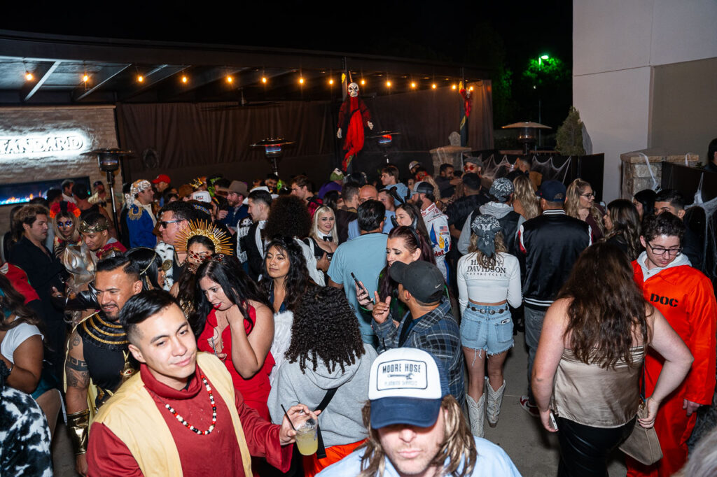 A large, packed crowd of people in various Halloween costumes at the outdoor patio area of The Standard in Fresno. Costumes visible include a man in a red and gold royal or tribal costume in the foreground, a woman dressed as Medusa (with a gold snake headpiece) in the center, and a person in a bright orange jumpsuit (possibly as an inmate) on the right. String lights hang overhead, and a large skeleton decoration is suspended from the back structure. The venue's "THE STANDARD" signage is visible on the far left next to an outdoor fireplace.
