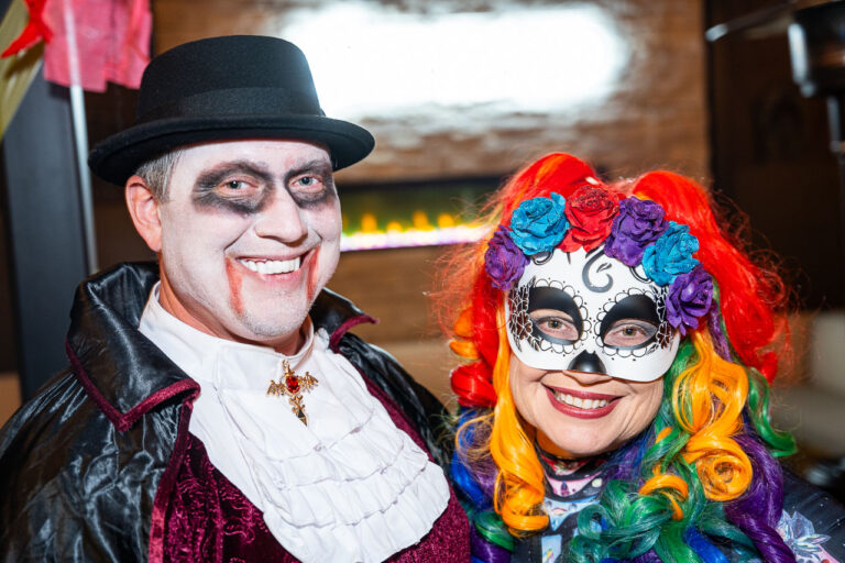 A smiling man and woman in elaborate costumes posing at The Standard in Fresno Halloween party. The man is dressed as a vampire, wearing a black top hat, a black and burgundy velvet cape, a ruffled white shirt, and a gold cross pendant. He has pale makeup with fake blood near his mouth. The woman is wearing a rainbow wig and a Day of the Dead (Sugar Skull) mask decorated with purple, blue, and red flowers.